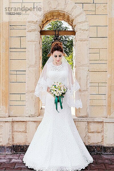 Eine schöne Braut im Brautkleid und mit einem Brautstrauß in den Händen steht im Freien vor einem Steinbogen mit einer Glocke. Das Konzept des Hochzeitsglücks. Hochzeitstag