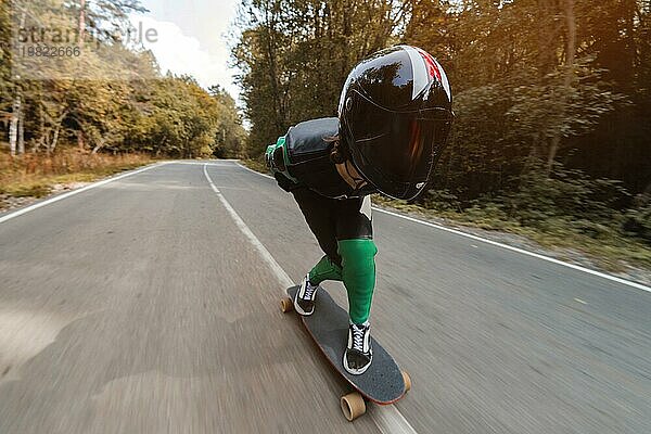 Ein junger Fahrer in einem speziellen Lederanzug und einem Ganzkörperhelm fährt mit hoher Geschwindigkeit auf seinem Longboard auf einer Landstraße im Herbstwald