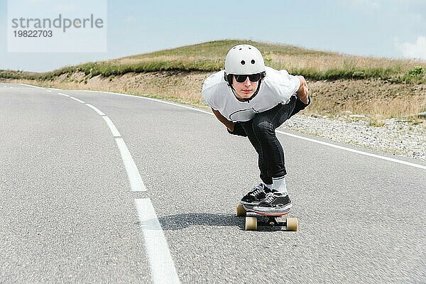 Der Typ mit dem Helm und der Sonnenbrille fährt mit seinem Longboard in einem speziellen Sportständer sehr schnell über die asphaltierte Straße