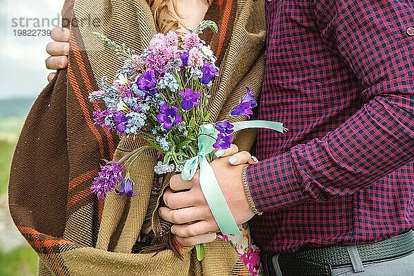 Nahaufnahme einer Hand  die einen Mann und seine Freundin mit Blumenstrauß umarmt. Kariertes Hemd und Strickpullover