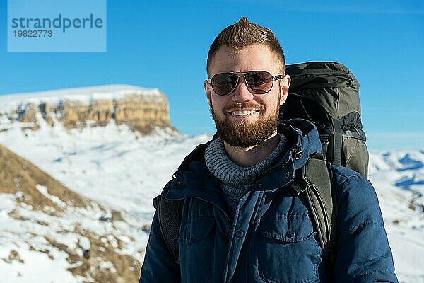 Porträt eines fröhlich lachenden Hipster Reisenden mit Bart und Sonnenbrille in der Natur. Ein Mann beim Wandern in den Bergen mit einem Rucksack und skandinavischen Wanderstöcken im Hintergrund einer Berglandschaft. Reisen Lebensstil Abenteuer Outdoor Erholung