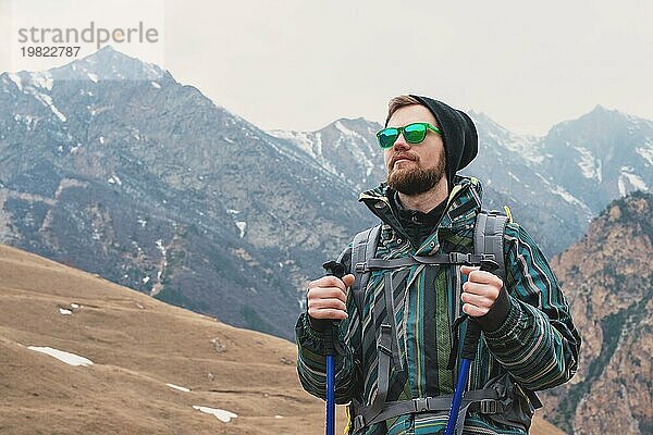 Ein Mann mit Bart und Sonnenbrille in einer Membranjacke  Hut  mit einem Rucksack und Stöcken für Nordic Walking  ein Reisender  der in der freien Natur steht und auf die Berge schaut