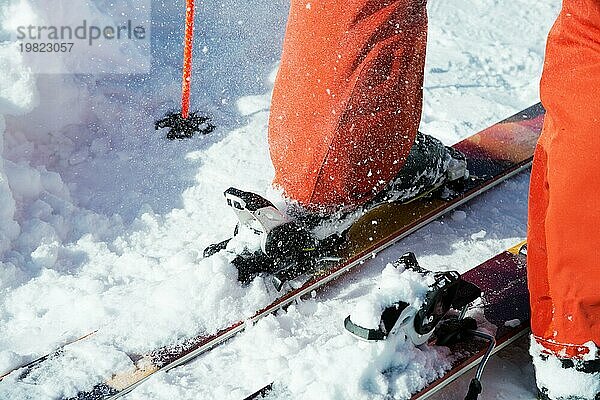 Orangefarbene Alpinskischuhe in einer Skihalterung. Ein Schuh ist vollständig auf den Skiern befestigt  der zweite nicht. Nahaufnahme. Orangefarbener Anzug