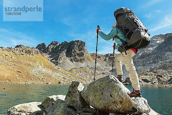 Schlankes und sympathisches Wandermädchen mit Sonnenbrille und blaür dünner Daunenjacke mit Rucksack und Trekkingstöcken steigt auf einen hohen Felsen vor dem Hintergrund von Felsen und einem hohen Bergsee