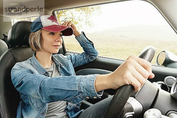 Glückliches hübsches Mädchen in Jeanskleidung und einer Mütze lächelnd am Steuer ihres Autos. Das Konzept der Verwirklichung des Traums vom Autofahren. Glück ist  ein Auto zu fahren. Freiheit