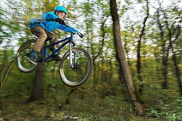 Ein junger Fahrer mit Helm und blauem Sweatshirt fliegt auf einem Fahrrad  nachdem er von einem hohen Kicker auf einem Waldradweg gesprungen ist. Aufnahme mit Langzeitbelichtung Verdrahtung