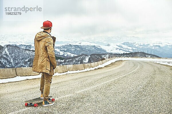 Stylish happy Junger Mann in Mütze und Hose Jogger rollen auf einem Longboard eine Bergstraße hinunter  genießen das Leben