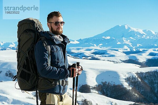 Ein Hipster Reisender mit Bart und Sonnenbrille in der Natur. Ein Mann wandern in den Bergen mit einem Rucksack und skandinavischen Wanderstöcke im Hintergrund eines Berges Elbrus Landschaft und blaün Himmel. Reisen Lifestyle Abenteuer Outdoor Erholung übertreffen