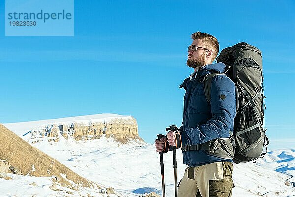 Ein Hipster Reisender mit Bart und Sonnenbrille in der Natur. Ein Mann wandert in den Bergen mit einem Rucksack und skandinavischen Wanderstöcken vor dem Hintergrund einer Berglandschaft und blauem Himmel. Reisen Lifestyle Abenteuer Outdoor Erholung Outdoor Sport