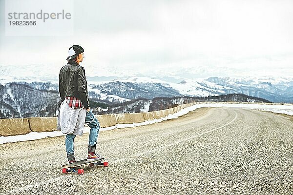 Stylische rothaarige Frauen in Jeansoveralls stehen mit einem Longboard auf einer Bodenwelle am Highway in den Bergen