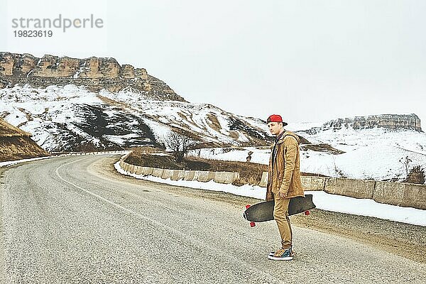 Stylish glücklich Junger Mann in einer Mütze und Hose Jogger stehend eine Bergstraße hinunter  ein Longboard in den Händen  das Leben genießen