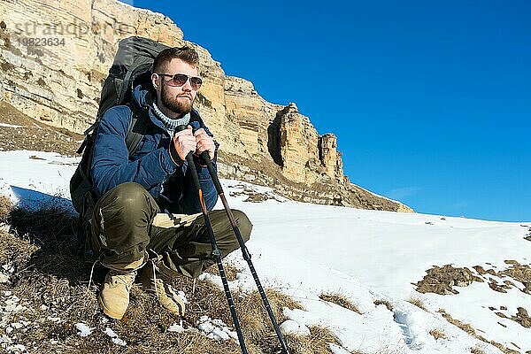 Porträt eines Hipster Reisenden mit Bart und Sonnenbrille sitzt in der Natur. Ein Mann wandert in den Bergen mit einem Rucksack und skandinavischen Wanderstöcken im Hintergrund einer Berglandschaft. Reisen Lifestyle Abenteuer im Freien Erholung im Freien spo