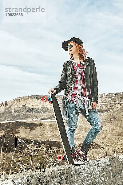 Stylische rothaarige Frauen in Jeansoveralls stehen mit einem Longboard auf einer Bodenwelle am Highway in den Bergen