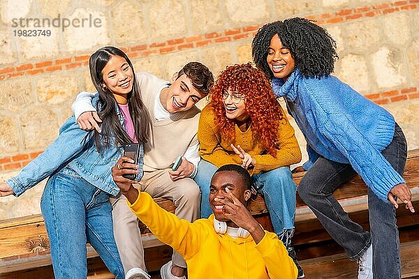 Coole  bunt gemischte Gruppe von TeenagerFreunden die zusammen auf einer Holzbank im Freien sitzen und ein Selfie machen