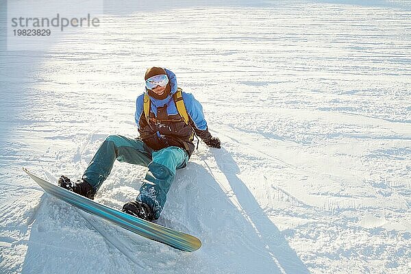 Ein lächelnder männlicher Snowboarder  der auf einem weißen Schnee sitzt. Das Konzept der Entspannung und Sport in den Bergen