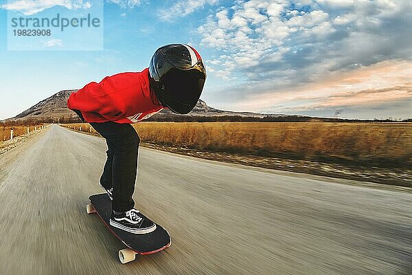 Ein junger Mann in einem Fullface Helm und einem Lederanzug in einem speziellen Gestell fährt ein Longboard auf afsaltu Sonnenuntergang im Hintergrund Berge und schönen Himmel mit hoher Geschwindigkeit