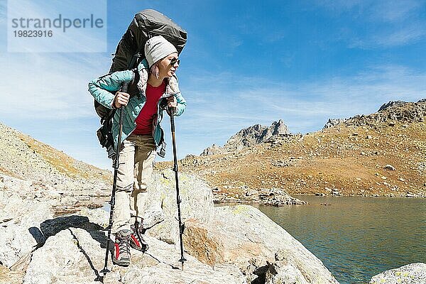 Schlankes und sympathisches Wandermädchen mit Sonnenbrille und blaür dünner Daunenjacke mit Rucksack und Trekkingstöcken steigt auf einen hohen Felsen vor dem Hintergrund von Felsen und einem hohen Bergsee