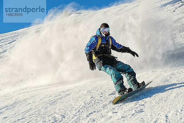 Ein Freeboard Snowboarder mit Skimaske und Rucksack fährt über die schneebedeckte Piste und hinterlässt einen Pulverschnee vor dem blaün Himmel