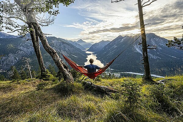Junger Mann sitzt in einer orangen Hängematte  Ausblick auf Plansee vom Schönjöchl  bei Sonnenuntergang mit Sonnenstern  Panoramablick auf Berge mit See  Plansee  Tirol  Österreich  Europa