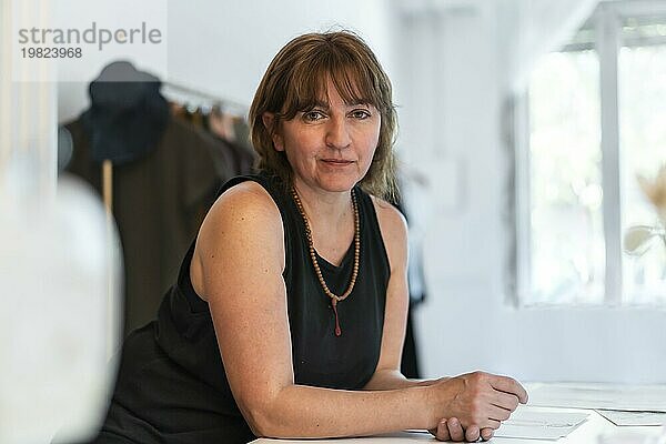 Eine Modedesignerin schaut in die Kamera  während sie sich in ihrem Atelier auf den Schreibtisch stützt. Kopierraum