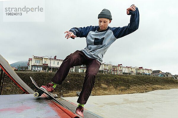 Ein jugendlicher Skateboarder mit Hut macht einen Rocks Trick auf einer Rampe in einem Skatepark vor einem bewölkten Himmel und einem Schlafplatz. Das Konzept des urbanen Stils im Sport