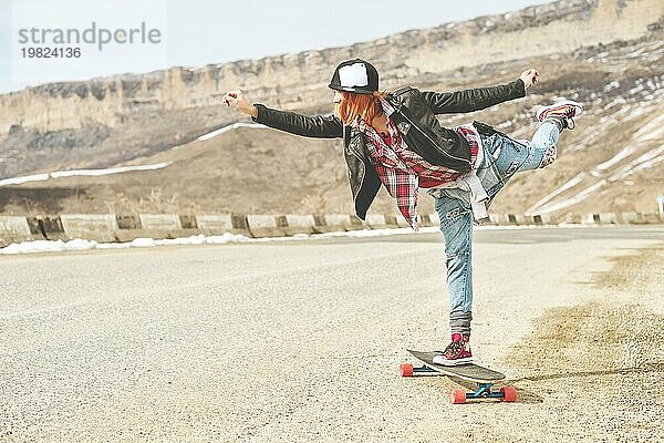 Stylish happy Junges Mädchen mit Mütze und Jeans Overall balanciert in der Mitte einer Bergstraße auf einem Longboard im Gleichgewicht