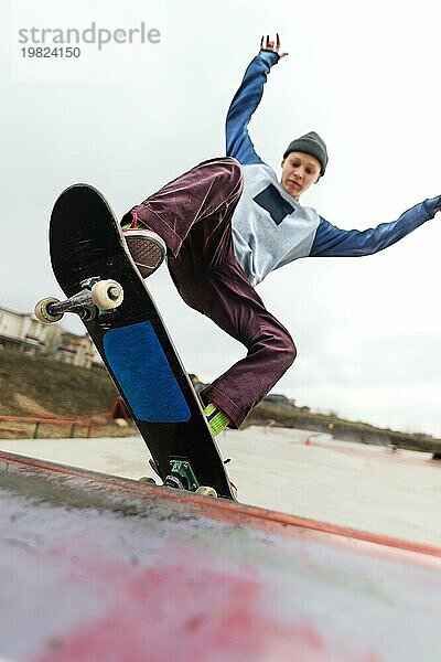 Ein jugendlicher Skateboarder mit Hut macht einen Rocks Trick auf einer Rampe in einem Skatepark vor einem bewölkten Himmel und einem Schlafplatz. Das Konzept des urbanen Stils im Sport