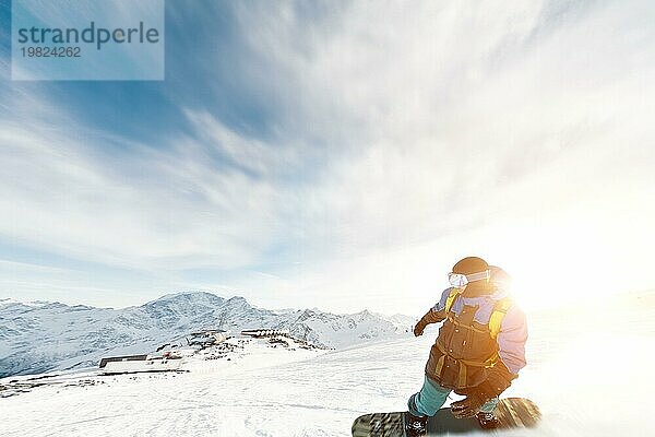 Ein Snowboarder mit Skimaske und Rucksack fährt auf einer schneebedeckten Piste und hinterlässt einen Pulverschnee vor dem blaün Himmel und der untergehenden Sonne. Foto in Bewegung