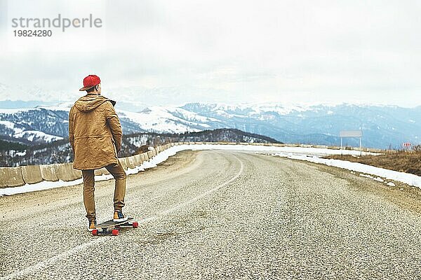 Stylish happy Junger Mann in Mütze und Hose Jogger rollen auf einem Longboard eine Bergstraße hinunter  genießen das Leben