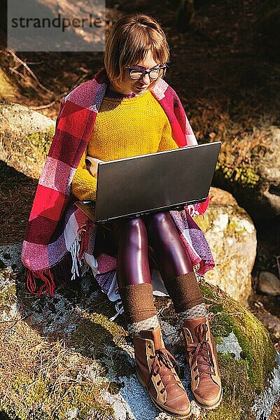 Ein getöntes Porträt eines lächelnden Freelancer Hipster Mädchens mit Brille  das in eine Decke gekleidet ist und mit einem Laptop auf einem Felsen in einem Nadelwald im warmen Sonnenlicht kniet. Das Konzept der freiberuflichen Arbeit in einem Abstand und Reisen ohne Unterbrechung w