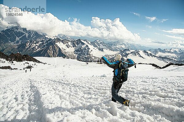 Müder Reisender mit Sonnenbrille und einem Rucksack  auf dem der Eispickel von einem schneebedeckten Gipfel herabhängt  vor dem Hintergrund schneebedeckter Berge und mit einem Stock für skandinavisches Wandern