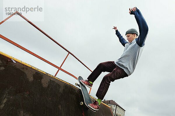 Ein jugendlicher Skateboarder mit Hut macht einen Rocks Trick auf einer Rampe in einem Skatepark vor einem bewölkten Himmel und einem Schlafplatz. Das Konzept des urbanen Stils im Sport