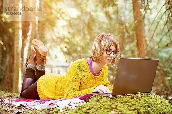 Toned Bild eines Freiberufler Mädchen Porträt in einem gelben Pullover und Brille Blick nachdenklich auf den Laptopbildschirm in der Natur in einem Nadelwald. Das Konzept der Freiberuflichkeit Freiheit für Reisen und das Fehlen der Bindung an das Büro. Ein Laptop liegt auf dem