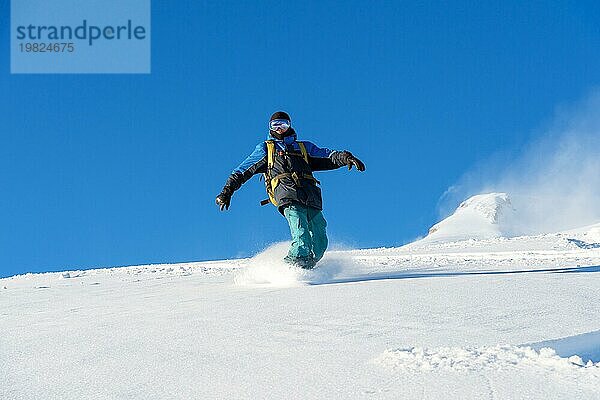 Ein Freeboard Snowboarder mit Skimaske und Rucksack fährt über die schneebedeckte Piste und hinterlässt einen Pulverschnee vor dem blaün Himmel