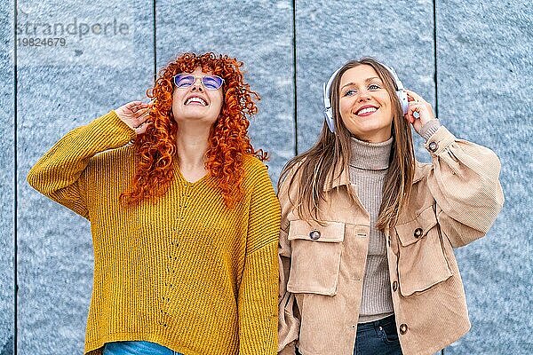 Frontalporträt von zwei glücklichen Frauen  die in der Stadt stehend mit Kopfhörern Musik hören