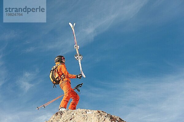 Ein Skisportler  der auf der Kante eines hohen Felsens steht  balanciert mit den Skiern in den Händen vor dem blaün Himmel und den Zirruswolken. Das Konzept des Skifahrens in den Bergen im Winter