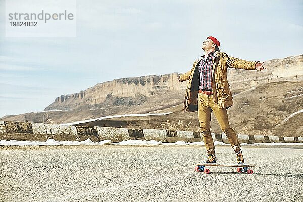 Stylish happy Junger Mann in Mütze und Hose Jogger rollen auf einem Longboard eine Bergstraße hinunter  genießen das Leben