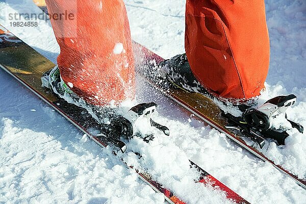 Orangefarbene Alpinskischuhe in einer Skihalterung. Ein Schuh ist vollständig auf den Skiern befestigt  der zweite nicht. Nahaufnahme. Orangefarbener Anzug