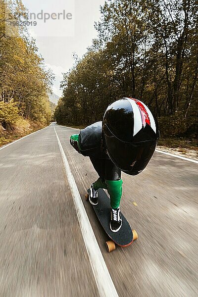 Ein junger Fahrer in einem speziellen Lederanzug und einem Ganzkörperhelm fährt mit hoher Geschwindigkeit auf seinem Longboard auf einer Landstraße im Herbstwald