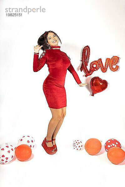 Junge Latina Mädchen in rotem Kleid und Schuhe springen lustig  schaut und lächelt in die Kamera  umgeben von Luftballons auf weißem Hintergrund mit Liebe Zeichen. Valentinstag Konzept