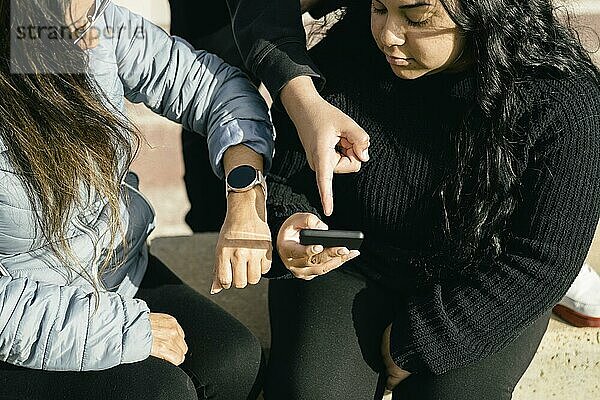 Glückliche Freunde  die mit ihren Smartphones und ihrer Smartwatch online Multimedia Inhalte austauschen  zwei schöne Latina Frauen  die im Freien sitzen