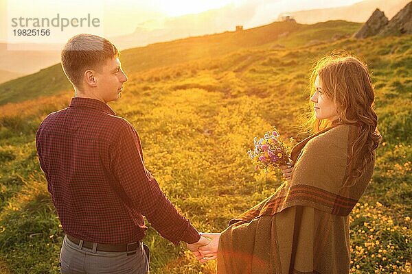 Ein junges Paar. Der Mann führt ein lockiges  in ein Plaid gehülltes Mädchen mit einem Blumenstrauß bei Sonnenuntergang. Das Konzept des Vertrauens und der glücklichen jungen Familie