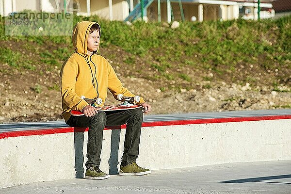 Teenager in Sweatshirt  Jeans und Turnschuhen  der in einem Skatepark sitzt  in die Ferne schaut und ein Skateboard hält