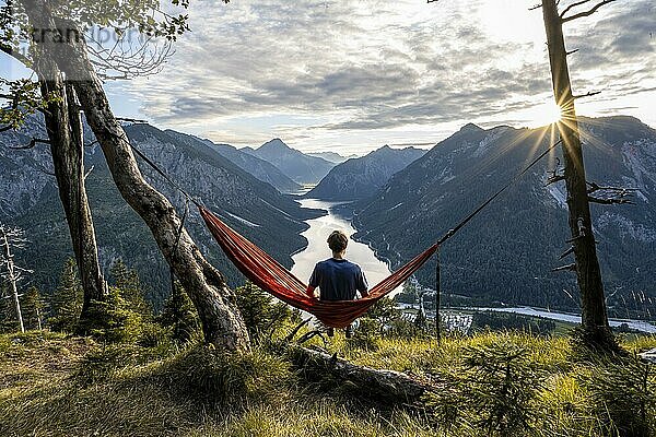 Junger Mann sitzt in einer orangen Hängematte  Ausblick auf Plansee vom Schönjöchl  bei Sonnenuntergang mit Sonnenstern  Panoramablick auf Berge mit See  Plansee  Tirol  Österreich  Europa