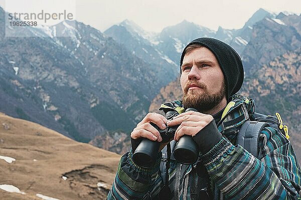 Ein Hipster Mann mit Bart  Hut  Jacke und Rucksack in den kaukasischen Bergen hält ein Fernglas  Abenteuer  Tourismus  Fährtensuche