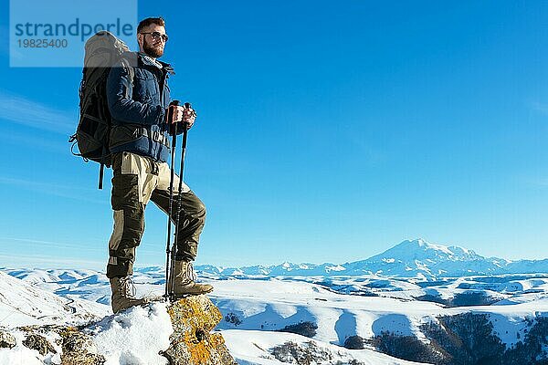 Ein Hipster Reisender mit Bart und Sonnenbrille in der Natur. Ein Mann wandern in den Bergen mit einem Rucksack und skandinavischen Wanderstöcke im Hintergrund eines Berges Elbrus Landschaft und blaün Himmel. Reisen Lifestyle Abenteuer Outdoor Erholung übertreffen