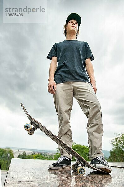 Ein junger langhaariger Skater im TShirt und mit einer langen Mütze steht mit einem Skateboard vor dem Hintergrund einer Stadtlandschaft