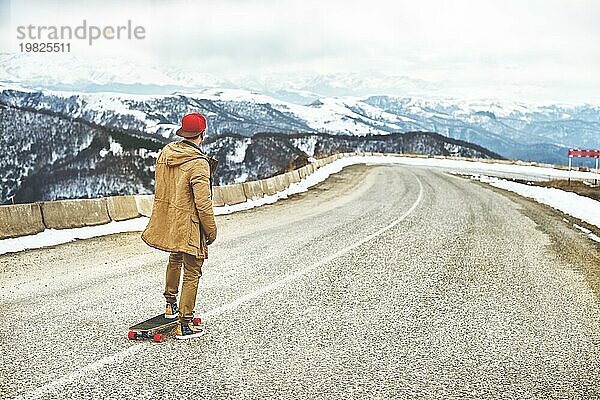 Stylish happy Junger Mann in Mütze und Hose Jogger rollen auf einem Longboard eine Bergstraße hinunter  genießen das Leben