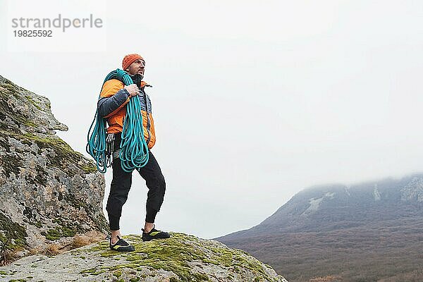 Voll ausgerüstet Der Hipster Kletterer steht in Daunenjacke und Bergschuhen auf einem hohen Felsen  mit einem Seil auf der Schulter und Karabinern am Gürtel. Das Konzept der extremen Ruhe in den Bergen