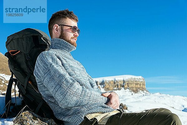 Porträt eines Hipsters mit Sonnenbrille und grauem Strickpullover vor dem Hintergrund einer schönen Winterlandschaft mit einem Tafelberg im Schnee. Das Konzept der Entspannung in den Bergen und Trekking mit Wandertouren